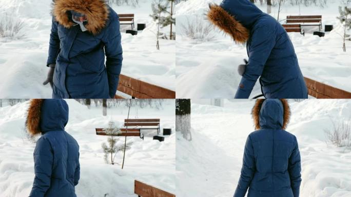 女人从长凳上站起来，甩掉积雪和树叶。冬季城市公园白天在下雪的天气下降雪。