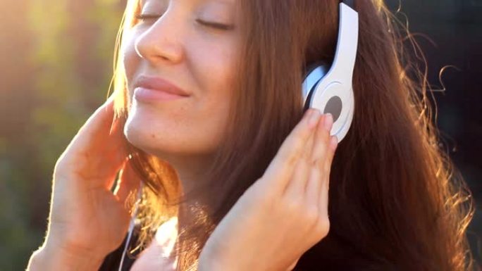 女人戴着耳机听音乐，闭着眼睛在户外日落时带着阳光。一个美丽的特写女孩的肖像。