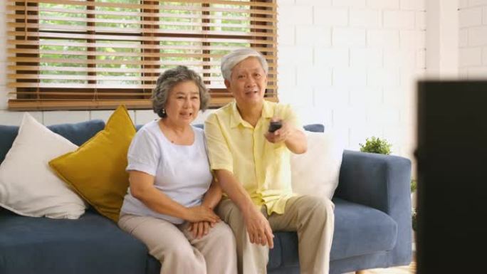亚洲夫妇老人坐在沙发上，用遥控器在家里的客厅换频道和看电视。从模糊的前景电视上平移