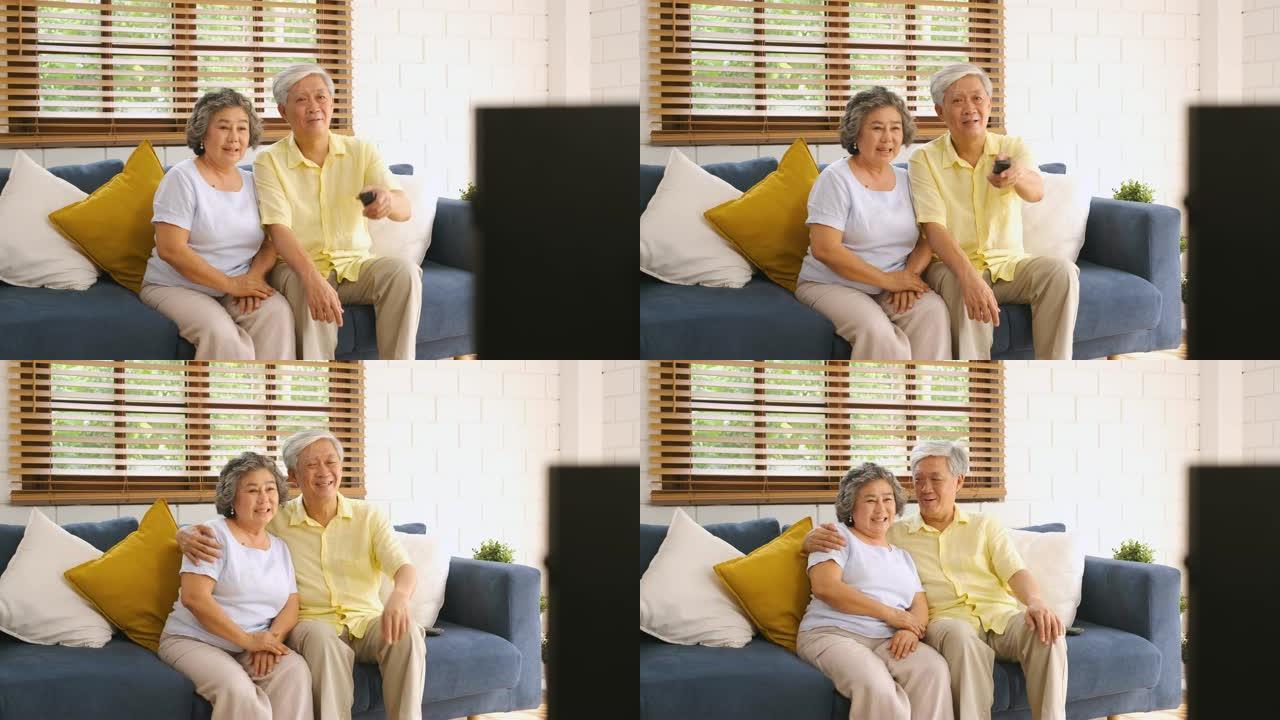 亚洲夫妇老人坐在沙发上，用遥控器在家里的客厅换频道和看电视。从模糊的前景电视上平移