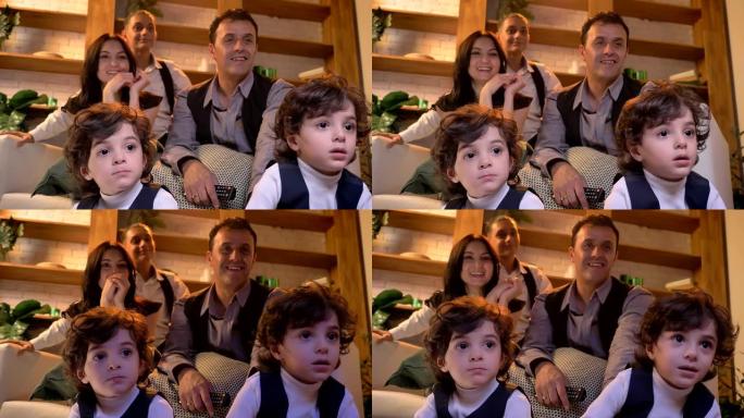 两个可爱的阿拉伯卷发双胞胎的肖像坐在客厅的地板上，他们的家人在背景上仔细地看电视。