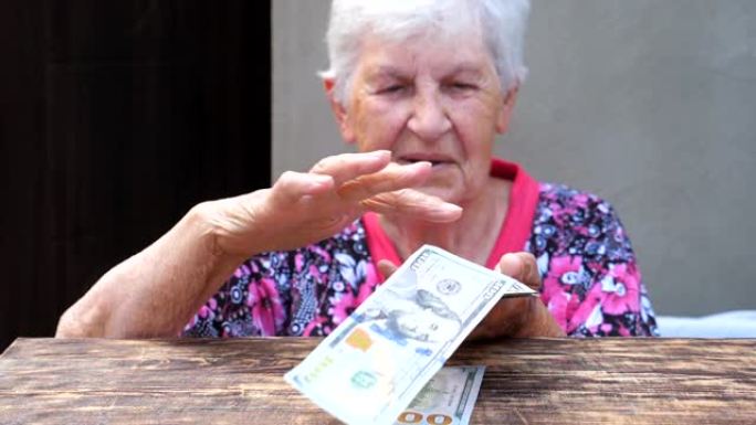 快乐的祖母手里拿着一捆钱，把外币撒在桌子上的肖像。老微笑的女人把100美元的钞票扔在桌子上。关闭慢动