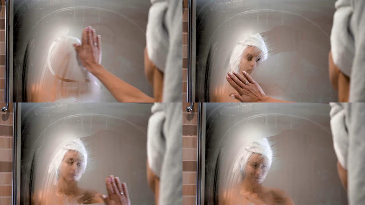 年轻的成年妇女在雾蒙蒙的浴室里擦拭镜子玻璃，沐浴后看着朦胧的反射。洗热水澡后，浴室里有浓稠的空气。女