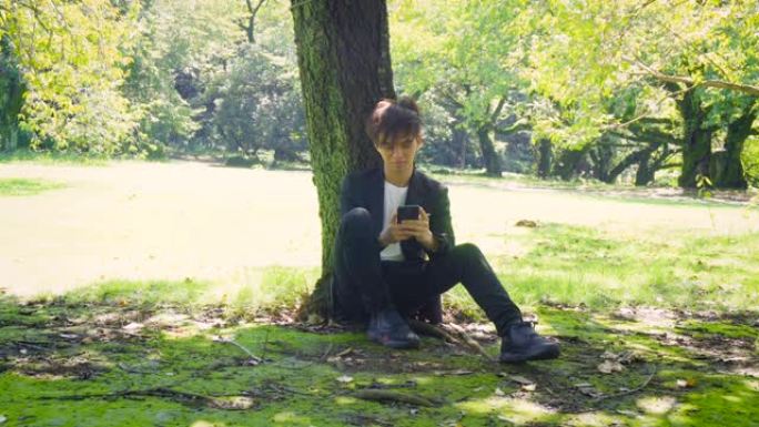 年轻的亚裔男子在树下使用手机-快乐的面部表情