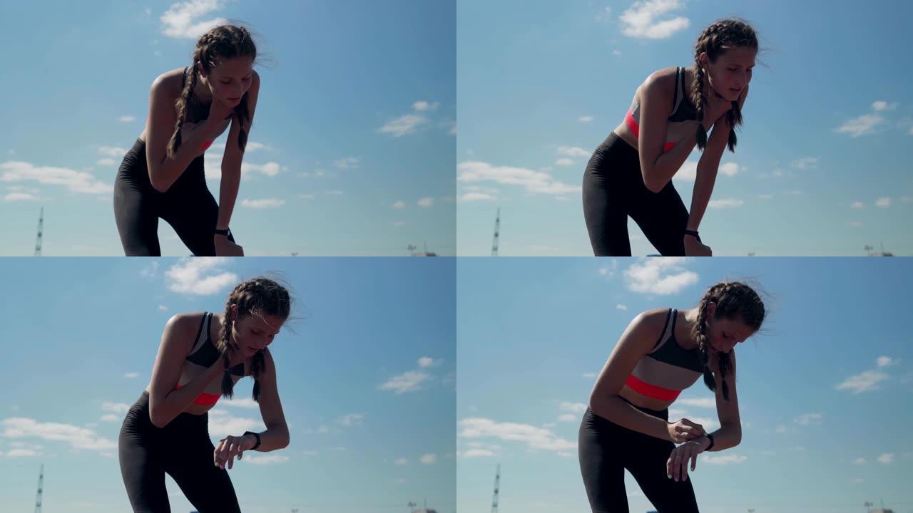 年轻的女运动员完成了跑步练习，弯腰呼吸并在蓝天背景下的健身追踪器上检查结果