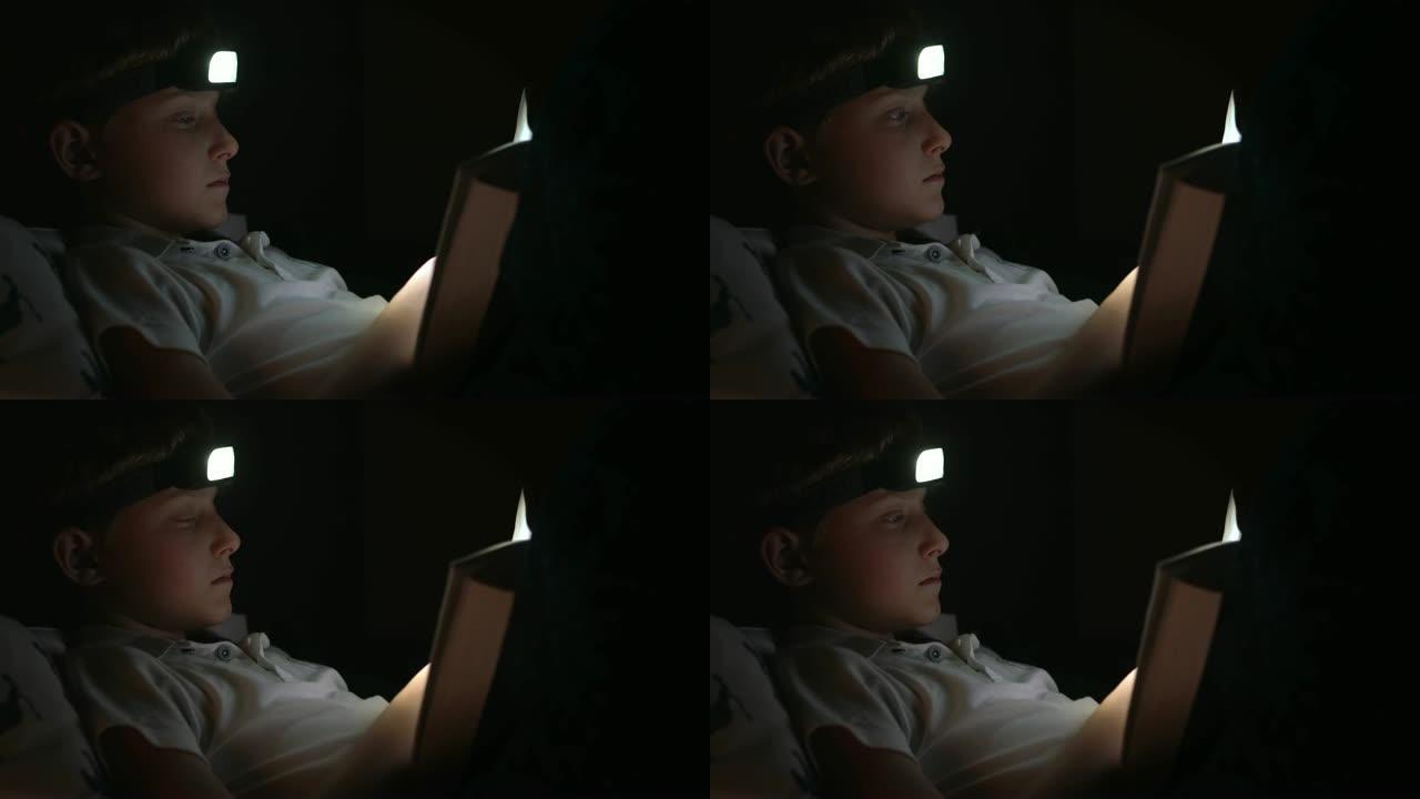 小读者男孩翻阅书页，他用头部火炬在床上看书。晚上在床上阅读概念照片。