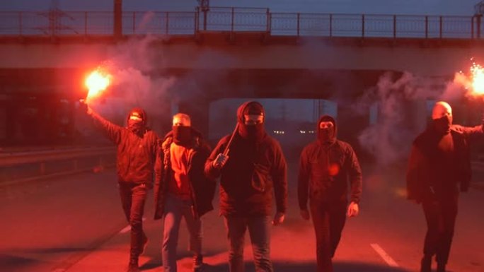 一群穿着巴拉克拉法帽的年轻人带着红色燃烧的信号弹在桥下的道路上行走，慢动作