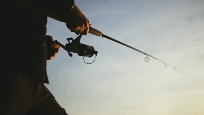 日落时分，渔夫在湖上钓鱼。一个穿着黑色背心和绿色帽子的渔夫用竹柄在纺纱上捕捉到掠食性鱼。
