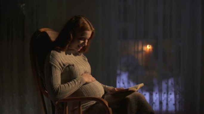 孕妇坐在黑暗房间的摇椅上。女士向未出生的婴儿大声朗读这本书