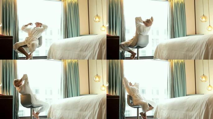 淋浴后，穿着白色浴袍的男人坐在酒店房间的旋转凳子上，光线充足的窗户前