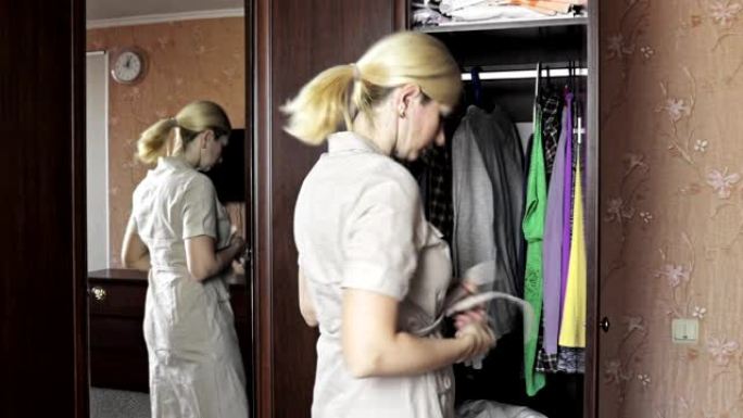 年轻的金发女子站在家里打开的衣柜前，解开她的家装，以便换衣服
