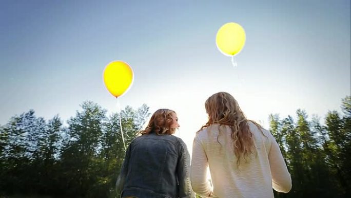 两个女孩带着气球穿过草地，一次旅行，另一个试图帮助，他们俩都笑了