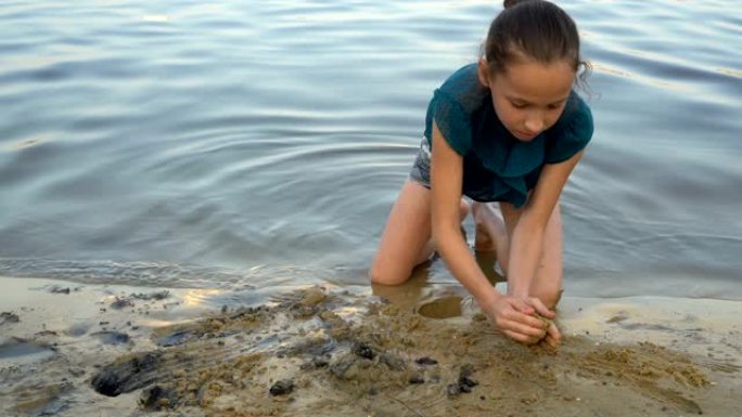 一个孩子，一个年轻的女孩，在日落时在河岸上建造一座沙堡。她坐在水边，用手耙沙。肖像。特写。4 k.2