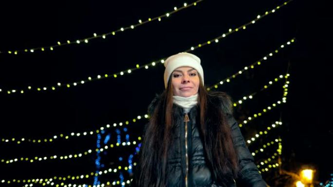 美丽的女孩微笑着在冬天穿过夜巷。街道上装饰着灯饰花环。摄像机的底视图。