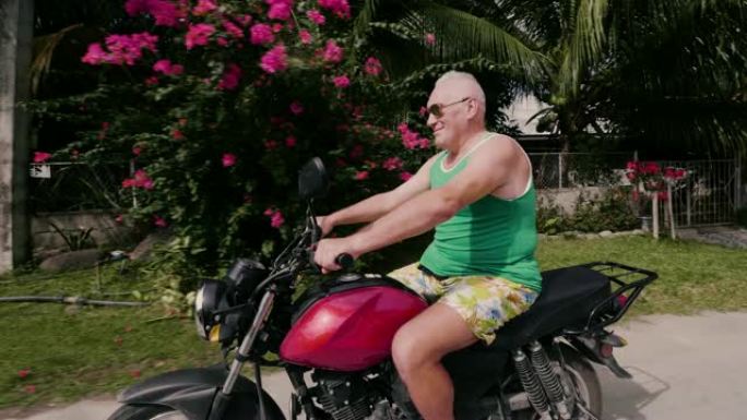 暑假期间在乡下开摩托车的老人。戴墨镜的高级男子在旅行时在热带村庄的街道上骑摩托车。
