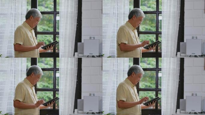 资深亚洲男子在家中站在窗前，每天的退休技术和生活方式时，会幸福地使用数字平板电脑