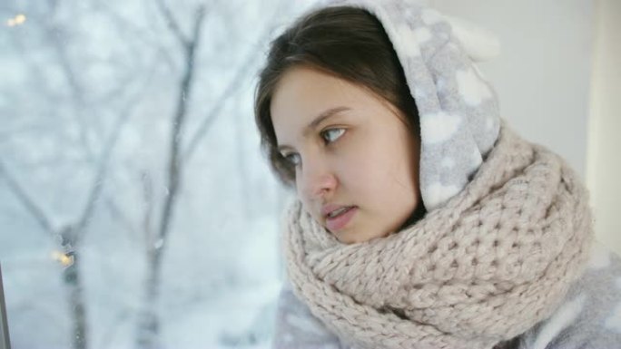 女孩少年冬天穿着温暖的睡衣和围巾坐在窗边，看起来就像窗外正在下雪。