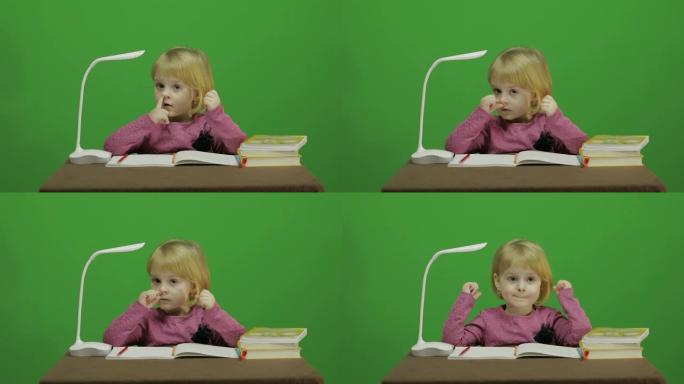 女孩坐在桌子旁，挑鼻子!课堂教育过程。快乐的三岁女孩。可爱的女孩微笑着。漂亮的小孩，3-4岁的金发女