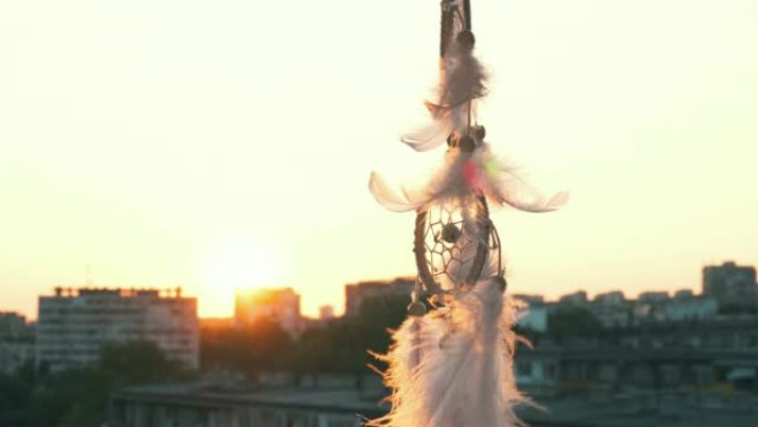 护身符梦想捕手在明亮的日落和城市灯光的背景下在微风中挥舞。白色淡淡的羽毛在风中升起。夕阳的圆盘。民族
