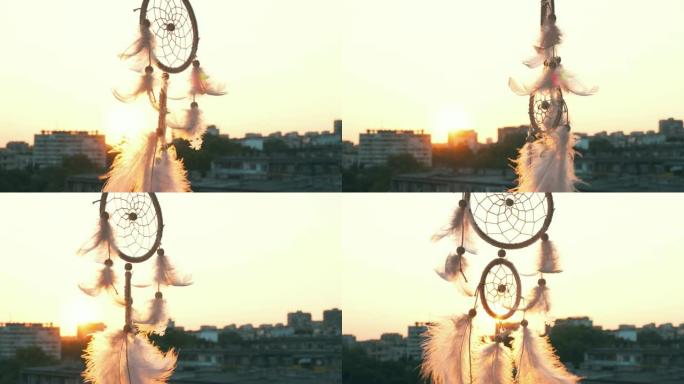 护身符梦想捕手在明亮的日落和城市灯光的背景下在微风中挥舞。白色淡淡的羽毛在风中升起。夕阳的圆盘。民族