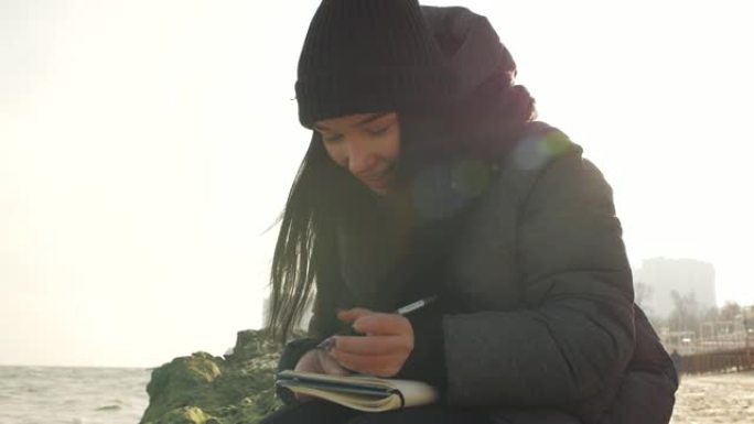 女孩微笑着准备用笔在笔记本上写字坐在海边的一块石头上