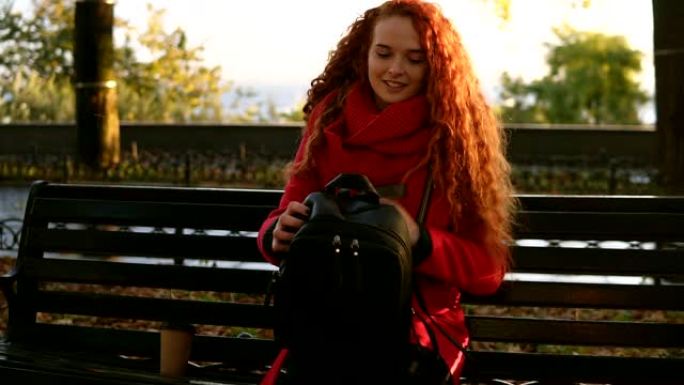 女人带着咖啡 “多哥” 和袋子在城市公园散步。卷发女士开始在公园的长凳上看书，在秋季公园休闲。穿着红