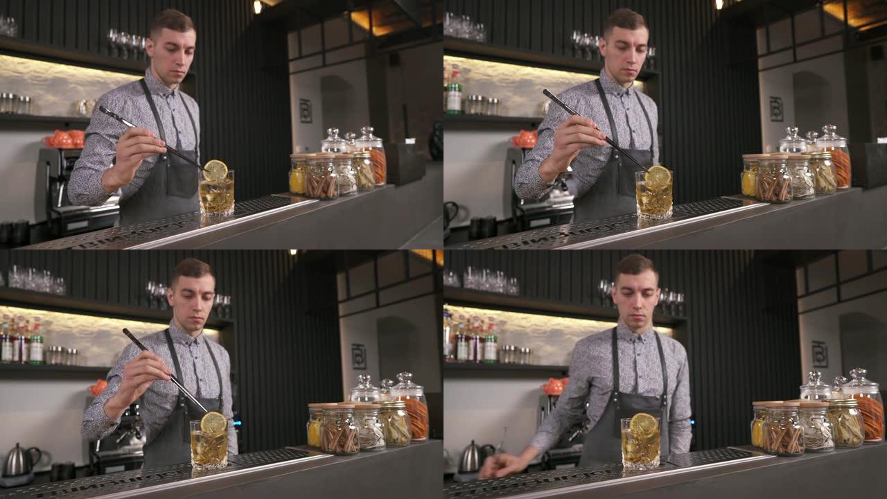 一位调酒师正在将一片柠檬和长镊子放入酒吧柜台上的一杯威士忌中。柜台后面站着一个酒吧标书