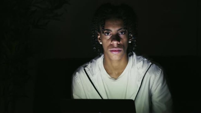 严肃的年轻人坐在黑暗房间的笔记本电脑上的肖像