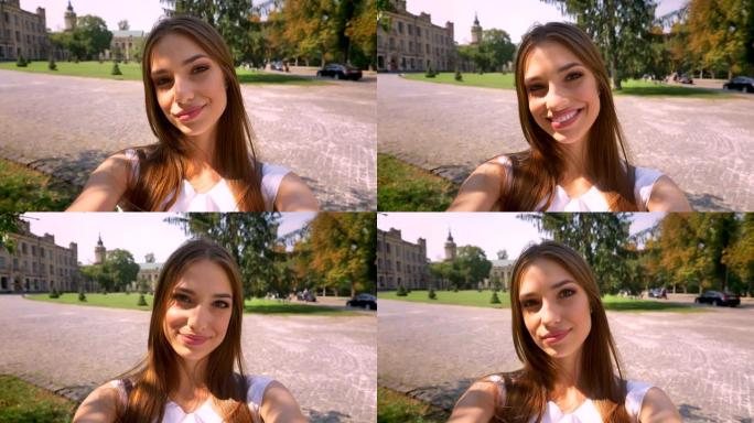 漂亮的年轻女子白天站在公园里，拿着相机，自拍，微笑，交流理念
