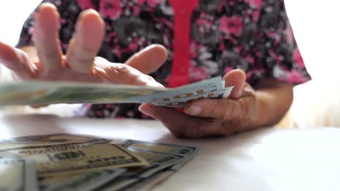 祖母手里拿着一捆钱，把100美元钞票扔在桌子上。老妇人的手臂将外币撒在前面的桌子上，对着镜头。关闭慢