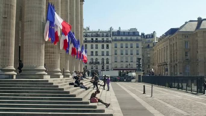 法国巴黎晴天，游客坐在万神殿前的楼梯上，法国国旗在柱子上挥舞。这是一个世俗的陵墓，里面有杰出的法国公
