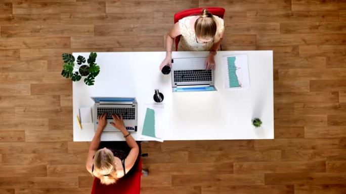 从上往下拍摄，两个辛勤工作的女性坐在木制工作室的桌子旁，在笔记本电脑上打字