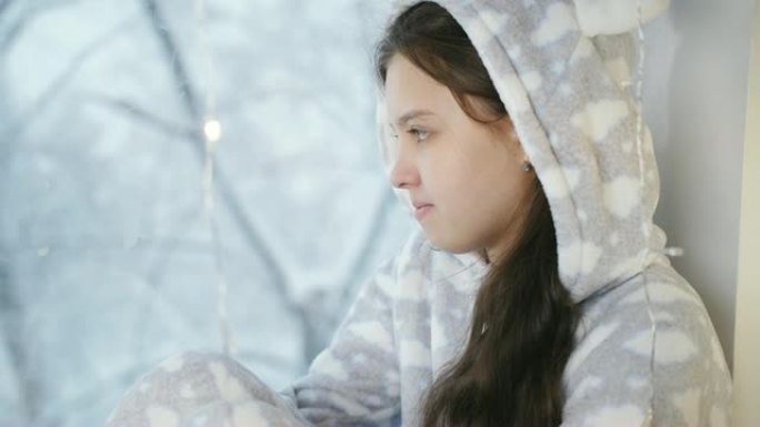 女孩少年冬天穿着温暖的睡衣和围巾坐在窗边，看起来就像窗外正在下雪。