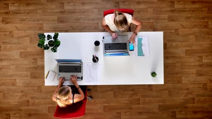 从上往下拍摄，两个金发碧眼的女人在木制的办公室里用他们的屏幕精确地检查纸质图表，蓝屏手机就放在笔记本