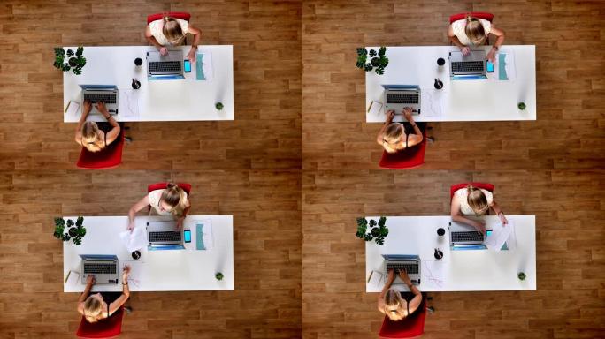 从上往下拍摄，两个金发碧眼的女人在木制的办公室里用他们的屏幕精确地检查纸质图表，蓝屏手机就放在笔记本