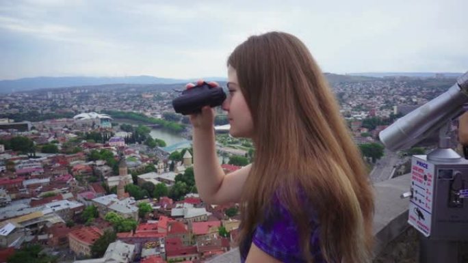 女孩年轻游客用双筒望远镜看着第比利斯美丽的城市景观背景
