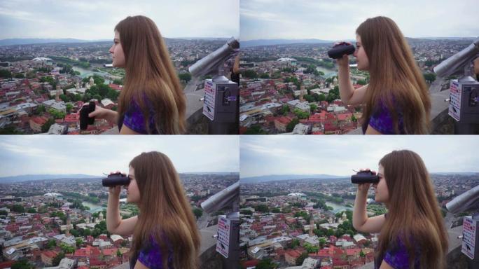 女孩年轻游客用双筒望远镜看着第比利斯美丽的城市景观背景