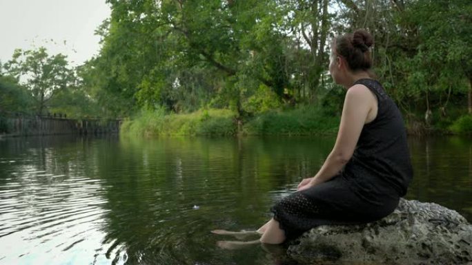 女人坐在溪流上的秋千上，在轻松的一天里，您的脚在水中缓慢而快乐地摆动。