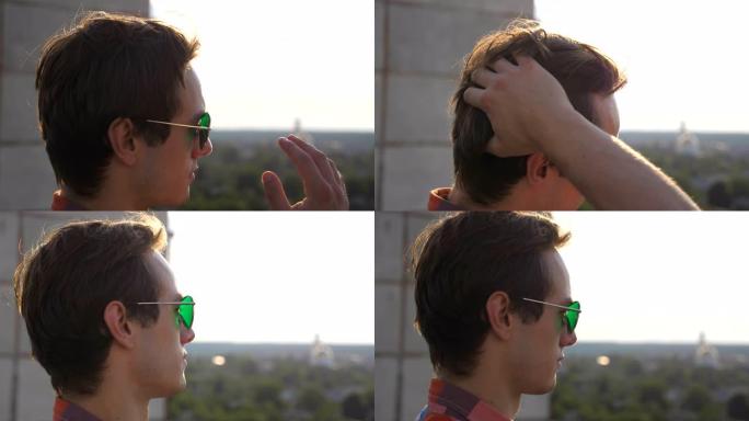 年轻男子的侧面观看全景城市从屋顶和直发。戴着墨镜的帅哥从屋顶上看城市风景，欣赏美丽的风景。侧视图特写