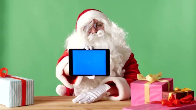 微笑的圣诞老人在相机中显示带有蓝屏的平板电脑，在桌子上显示礼物，在背景中显示绿色chromakey。