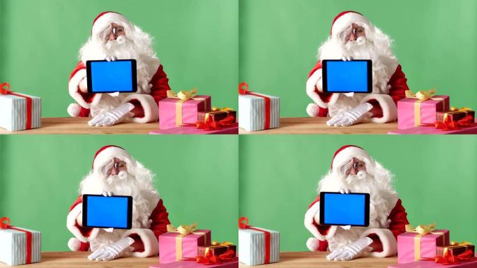 微笑的圣诞老人在相机中显示带有蓝屏的平板电脑，在桌子上显示礼物，在背景中显示绿色chromakey。