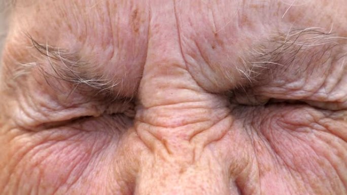 成熟女人的肖像拧起灰色的眼睛，然后睁开。关闭老祖母皱着皱纹的脸，带着悲伤的景象看着镜头。高级女士的悲