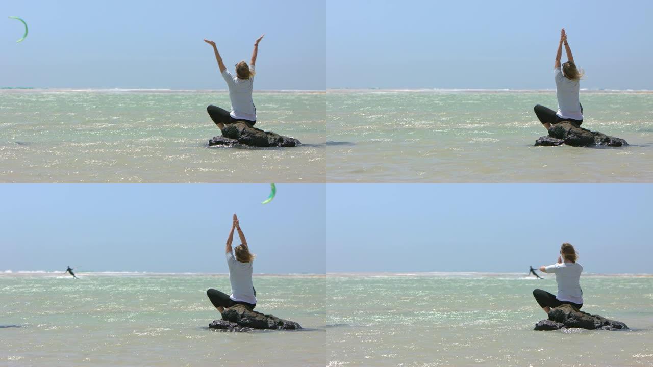 迷人的金发女人在平坦的水中做瑜伽练习，而她的丈夫正在风筝冲浪