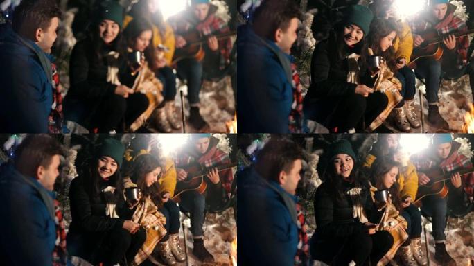 一群快乐的朋友坐在火炉边的冬季森林里，吃棉花糖。一个年轻女子调情和微笑。