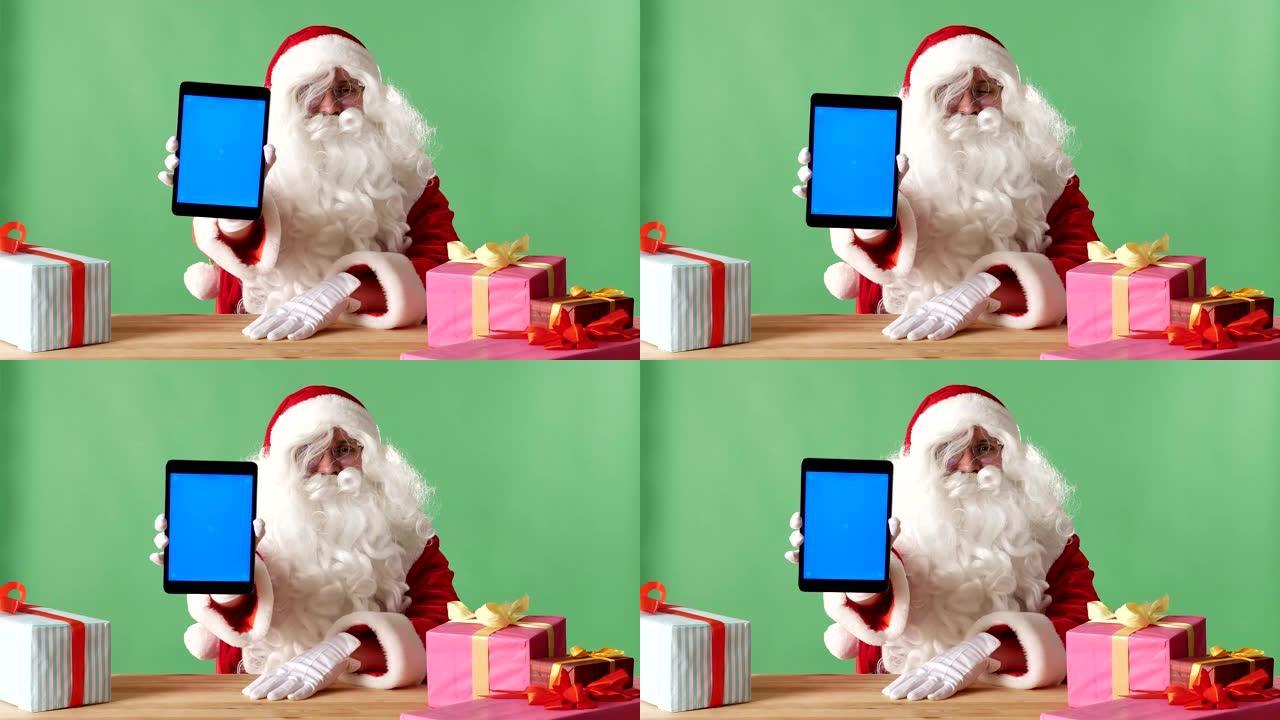 微笑的圣诞老人在相机中显示带有蓝屏的平板电脑，在桌子上显示礼物，在背景中显示chromakey。