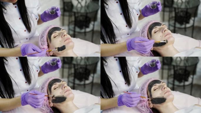医生-美容师在美容中心用刷子涂抹黑色面膜，以清洁和滋润面部皮肤。