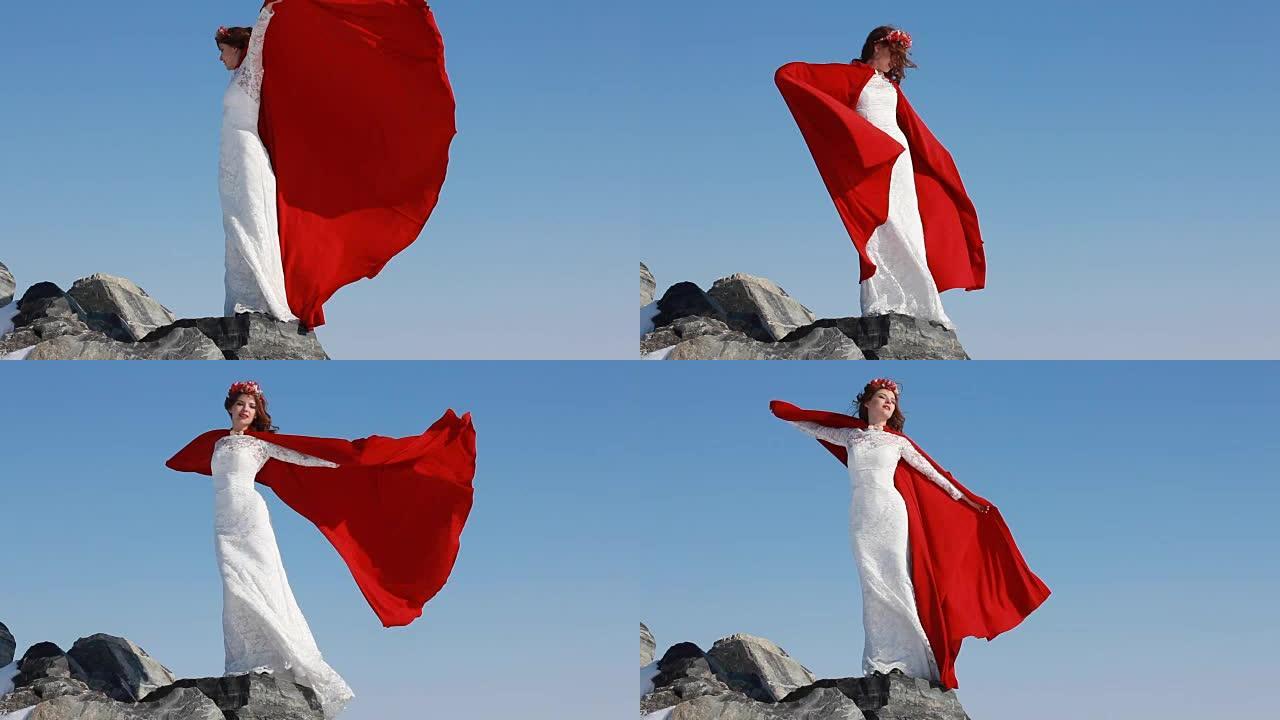 新娘站在悬崖上，在蓝天和冬天的背景下，风吹着红色的雨衣。