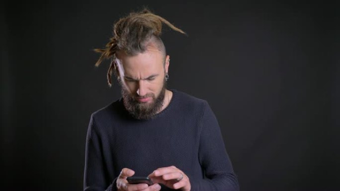 怪异的高加索人的肖像，留着辫子，在黑色背景上玩乐地刺入智能手机。