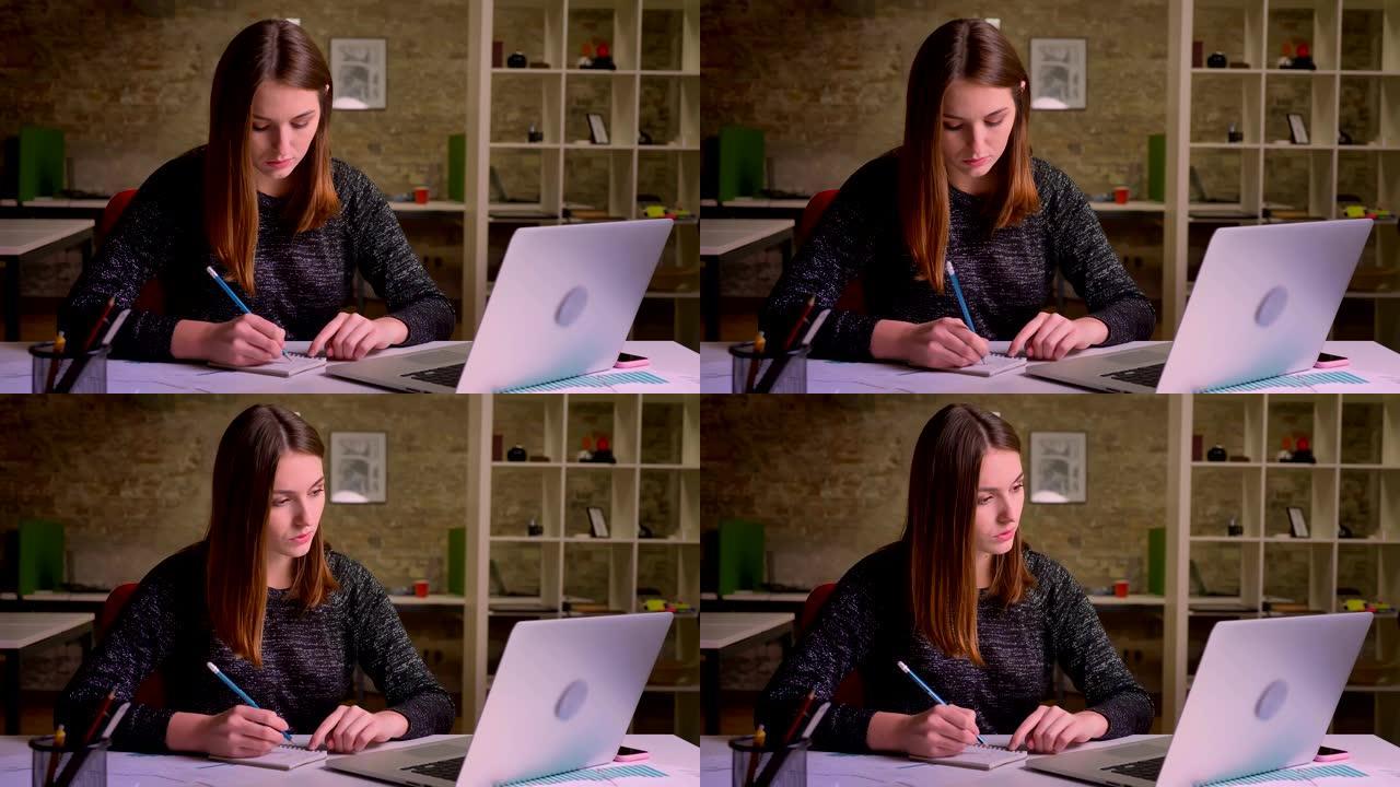 年轻的红发女人在笔记本电脑前工作，在办公室里用养老金和笔记本做笔记