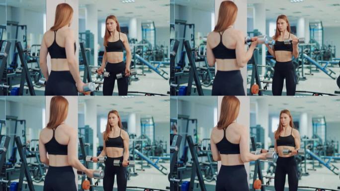 一个穿着黑色紧身裤和上衣的美女正在健身房用哑铃对着镜子做二头肌运动。